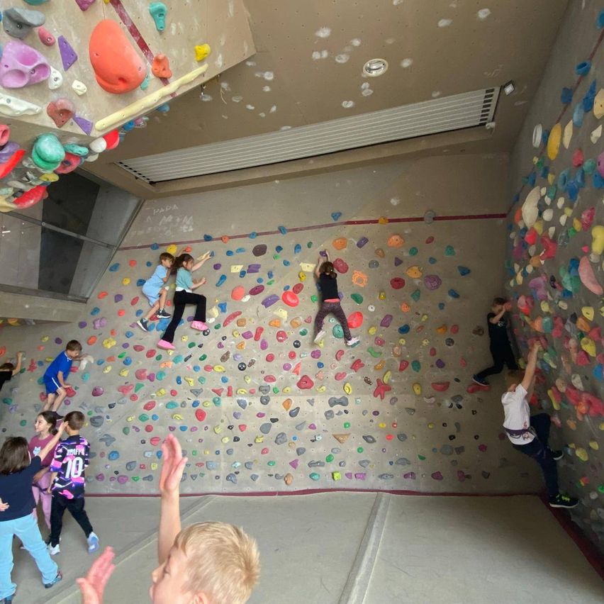 Kinder klettern an einer Wand mit verschiedenen Formen und Farben von Griffen. Einige sind oben, andere sind halb oben. Sie tragen Turnschuhe. Die Wand ist mit unterschiedlich gefärbten Griffen bedeckt.