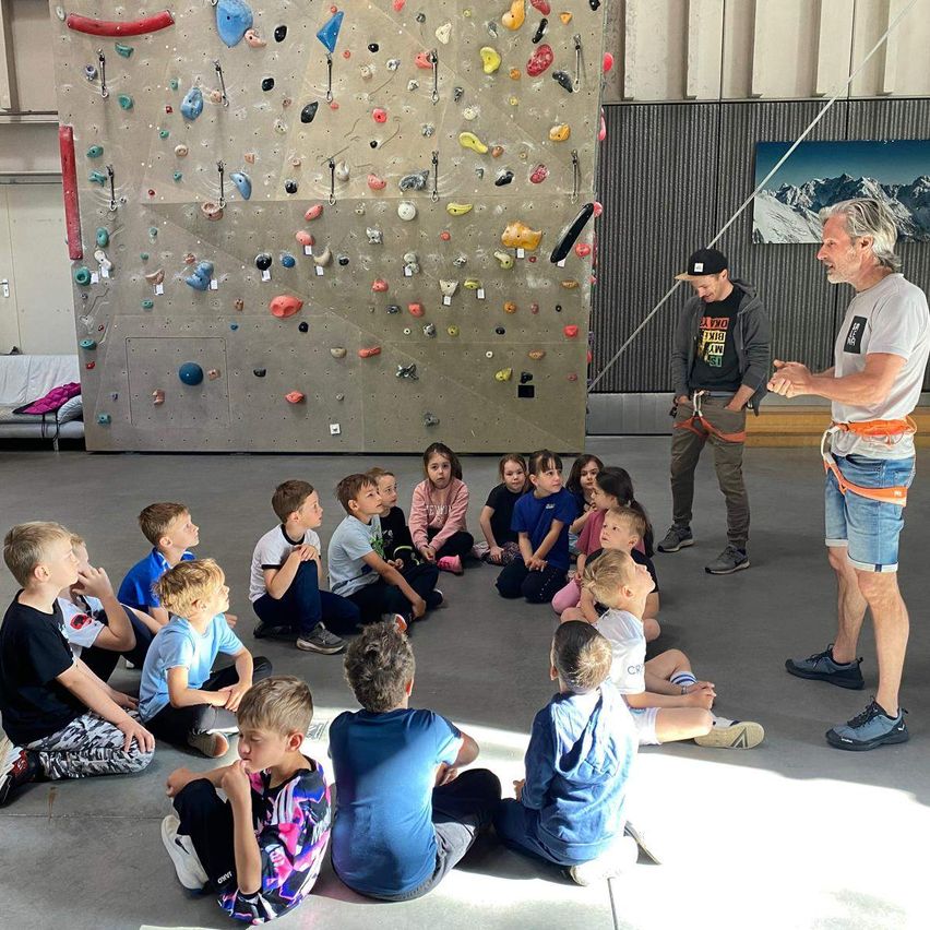 In einer Indoor-Kletteranlage spricht ein Mann zu einer Gruppe von Kindern, die im Kreis um ihn herum sitzen. Die Wand hinter ihnen ist mit verschiedenen Klettergriffen bedeckt.