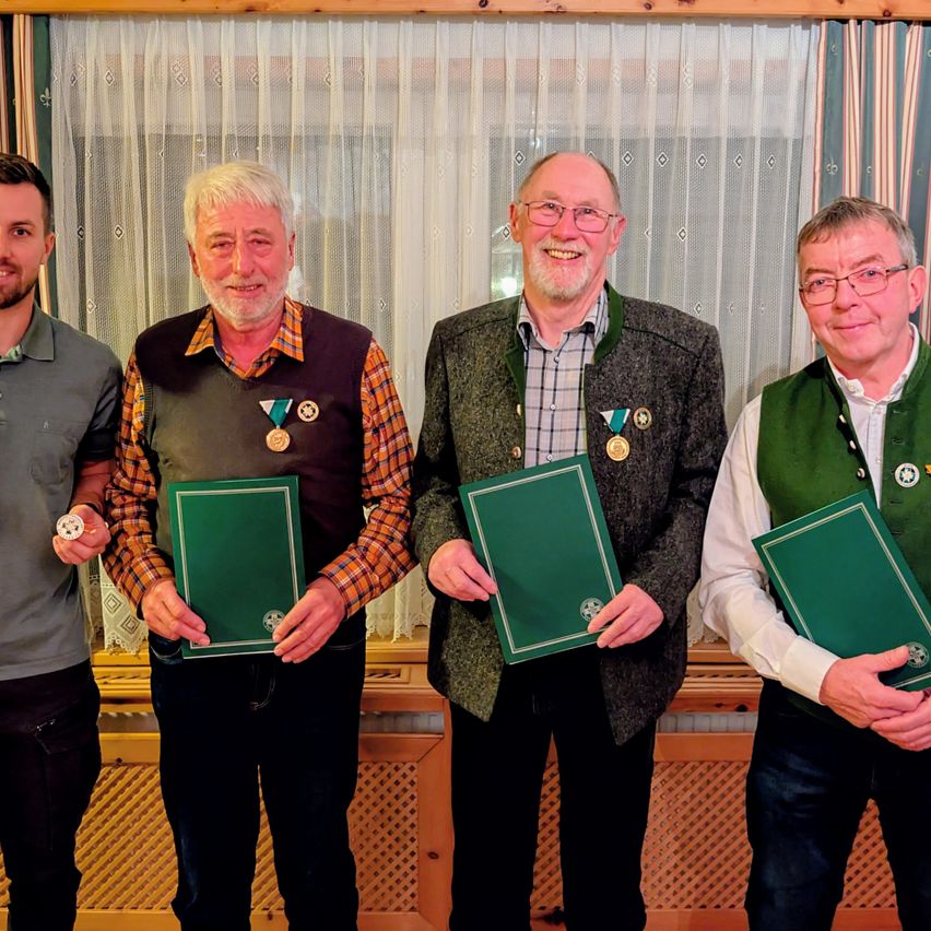Vier Männer stehen vor einem Vorhang und halten grüne Ordner in den Händen. Jeder trägt Medaillen um den Hals. Der Mann ganz links trägt ein graues Polo-Shirt.