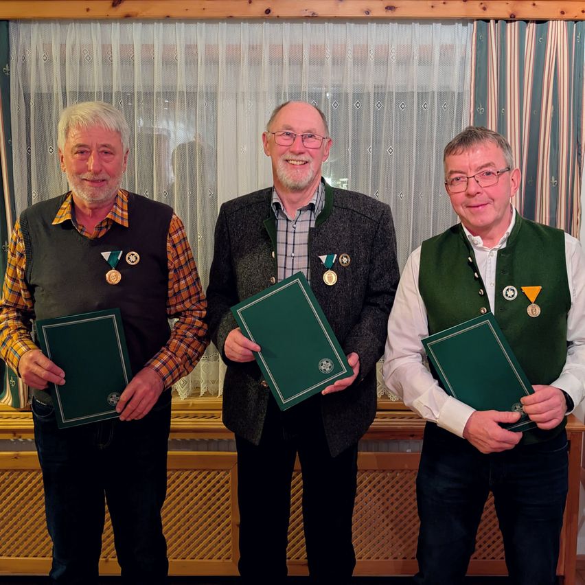 Drei Männer stehen zusammen, lächelnd, und halten grüne Ordner. Sie tragen Medaillen und Westen, möglicherweise bei einer Feier oder Preisverleihung. Ein Vorhang mit Diamantmuster befindet sich hinter ihnen.