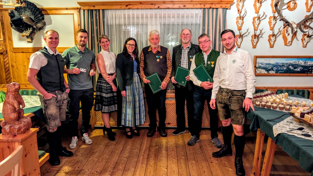 Eine Gruppe von Menschen steht in einem Raum und hält grüne Dokumente. Sie tragen verschiedene Kleidungsstücke, einige mit Medaillen um den Hals. Ein Mann in Lederhosen hält eine Karte. Hinter ihnen befindet sich ein weißer Vorhang und eine Holzwand.