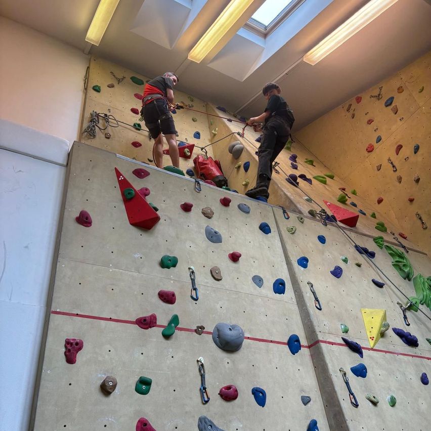 Zwei Personen klettern eine Indoor-Kletterwand mit verschiedenen farbigen Griffen und Haltegriffen. Ein Kletterer ist oben, während der andere aufsteigt, beide mit Seilen gesichert. Die Wand ist hell beleuchtet.
