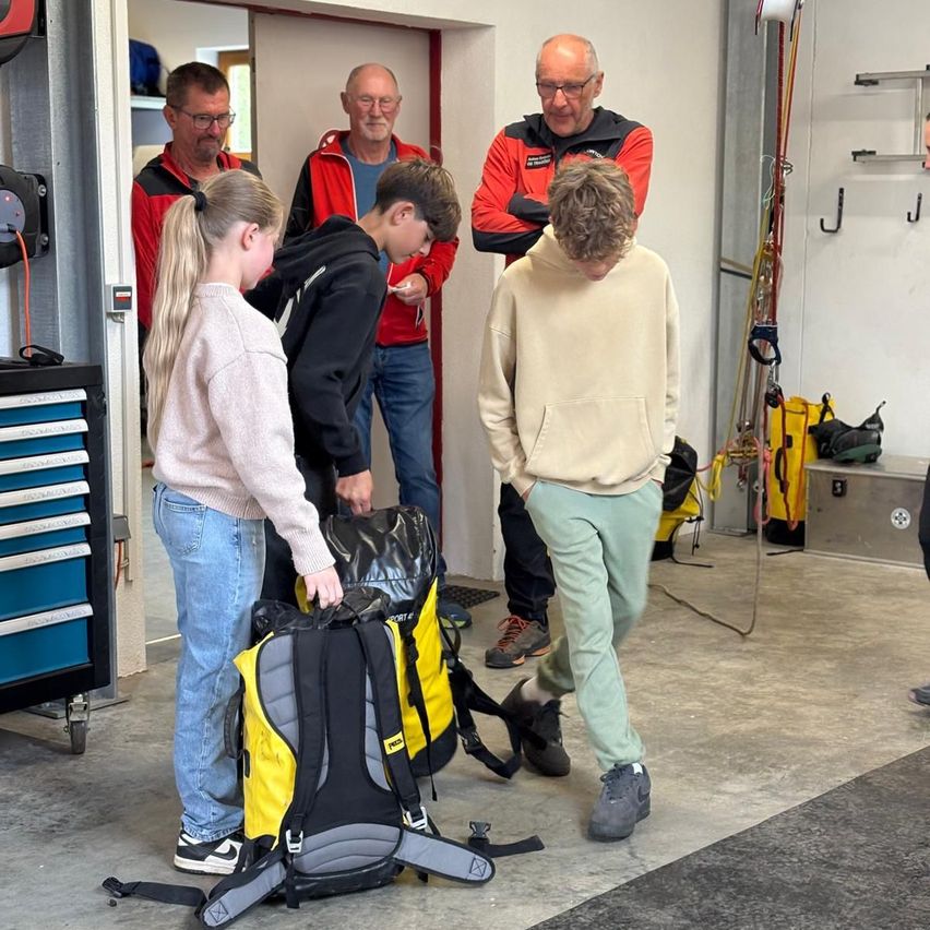 Eine Gruppe von Menschen versammelt sich in einer Garage. Eine Frau und ein Mann untersuchen einen Rucksack. In der Nähe stehen zwei Männer und beobachten. Ausrüstung und Werkzeuge hängen an den Wänden.