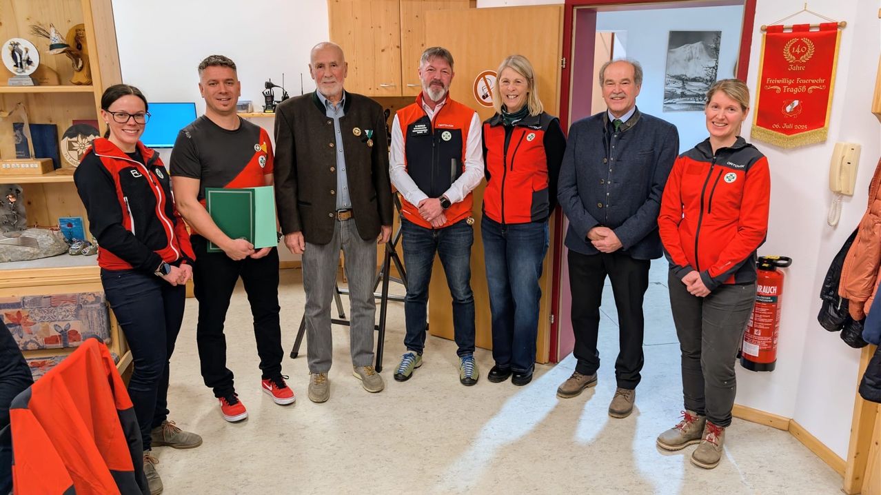 Five people stand in a room, smiling. The man on the left holds a green folder. Another man holds a folder. Behind them, a wooden cabinet is placed against the wall.
