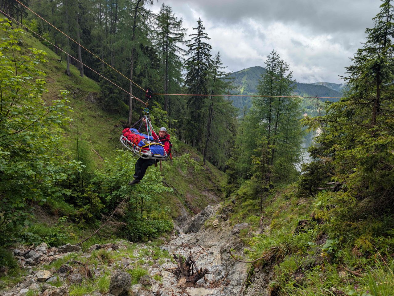 Ein Retter zieht eine Trage mit einer Person daran durch einen Wald mit einer Seilwinde. Die Person trägt einen roten und blauen Mantel und einen Helm. Der Wald ist von Bergen umgeben.