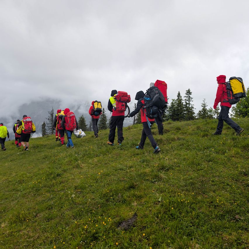 Eine Gruppe von Wanderern, die rote und gelbe Jacken tragen und Rucksäcke tragen, geht unter bewölktem Himmel in einem Grasbereich bergauf.