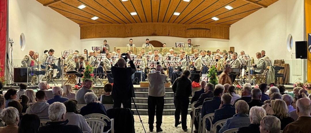 Eine große Gruppe von Musikern tritt auf der Bühne vor einem Publikum auf, das in Stühlen sitzt. Die Bühne ist mit Musikinstrumenten und Blumen geschmückt. Das Publikum sitzt in Reihen auf gepflastertem Boden.