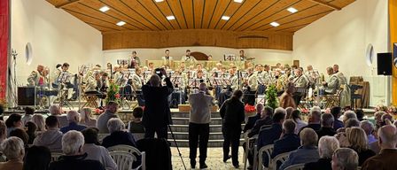 Eine große Gruppe von Musikern tritt auf der Bühne vor einem Publikum auf, das in Stühlen sitzt. Die Bühne ist mit Musikinstrumenten und Blumen geschmückt. Das Publikum sitzt in Reihen auf gepflastertem Boden.