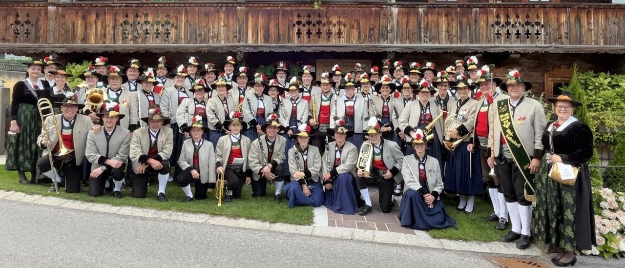 Eine Gruppe von Musikern in traditioneller bayerischer Kleidung, einschließlich Hüten und Uniformen, posiert für ein Foto vor einem Holzgebäude mit Balkonen.