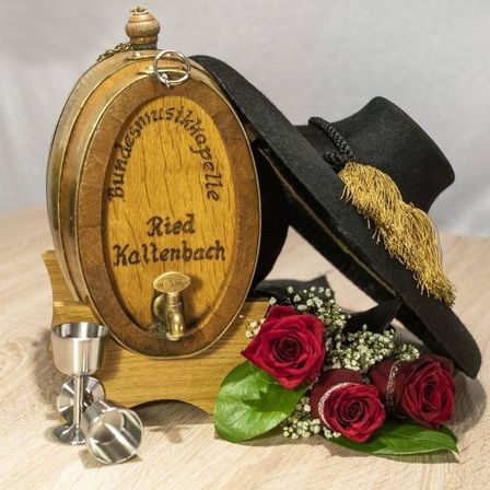 Ein hölzerne Tonne mit dem Text Ried Kaltenback steht auf einem Holzständer. Ein schwarzer Hut, ein silberner Becher, ein Strauß Rosen und einige grüne Blätter sind um die Tonne herum platziert.