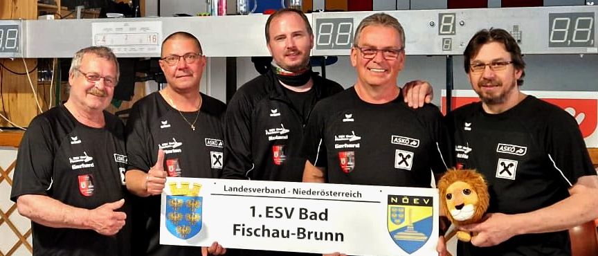 Fünf Männer stehen in einer Reihe, lächeln und halten ein großes Banner mit der Aufschrift '1. ESV Bad Fischau-Brunn'. Sie tragen alle passende schwarze Shirts. Der Mann auf der linken Seite hält ein Plüschtier. Sie stehen vor einer Holzbank.