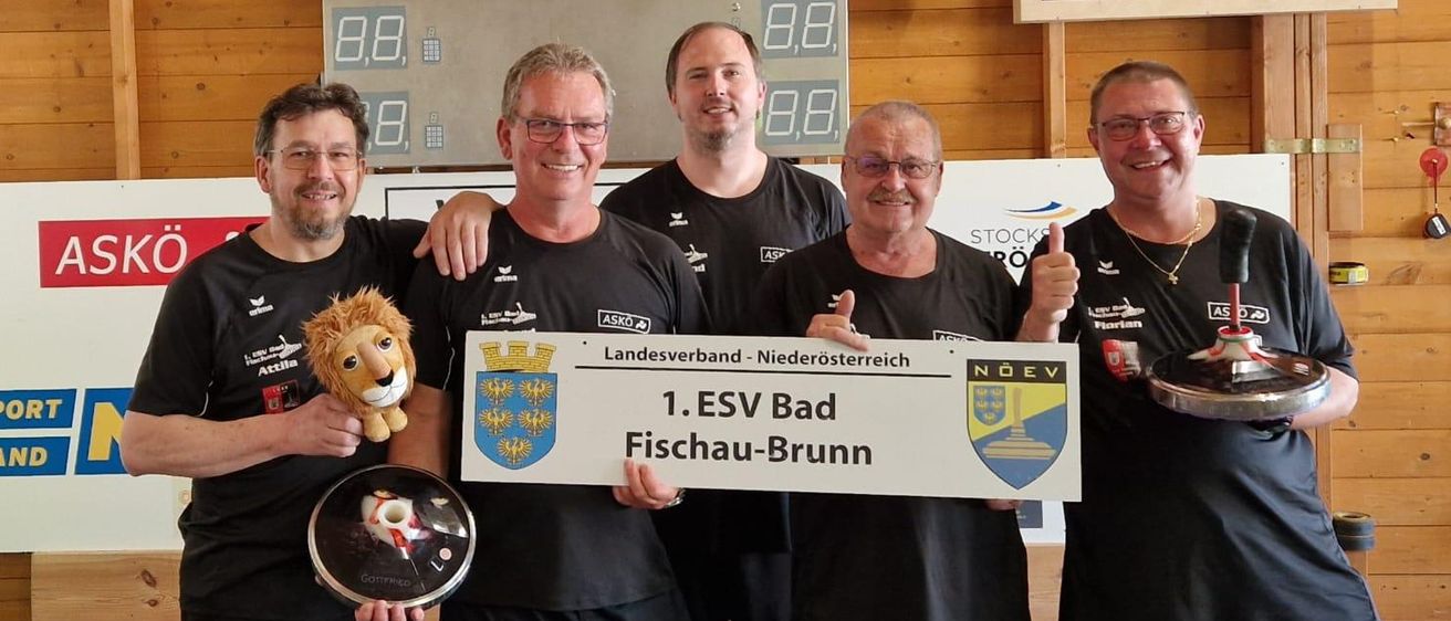 Fünf Männer posieren für ein Foto mit Pokalen und einem Zertifikat in einer Sporthalle. Sie tragen passende schwarze Shirts und Shorts. Ein Löwenkuscheltier und Spielteile sind in ihren Händen. Hinter ihnen zeigt eine Anzeigetafel Zahlen an.