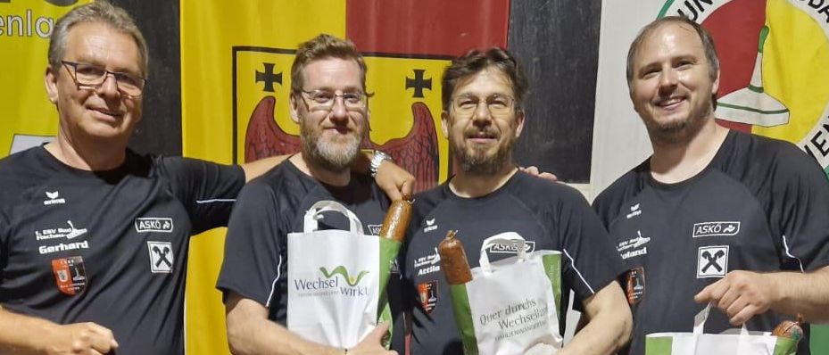 Vier Männer posieren für ein Foto, alle in gleichen schwarzen T-Shirts. Sie halten Tüten und Würste, was auf eine Feier hindeutet. Dahinter ist ein bunter Hintergrund mit Fahnen und Logos.