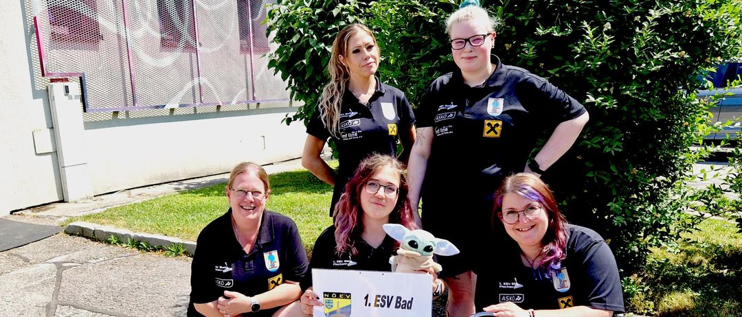 Fünf Frauen in schwarzen Uniformen mit Aufnähern posieren für ein Foto. Die Frau in der Mitte hält ein Schild mit der Aufschrift '1. ESV Bad Fischau Brunn' und ein Plüschtier. Sie befinden sich auf einer gepflasterten Oberfläche mit Pflanzen und einem Zaun im Hintergrund.