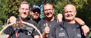 Vier Männer in schwarzer Sportbekleidung halten einen Korb voller Snacks. Ein Mann trägt eine Kappe und eine Brille, ein anderer zeigt den Daumen hoch. Sie stehen in einem Außenbereich mit Bäumen und einer Wasserrutsche im Hintergrund.