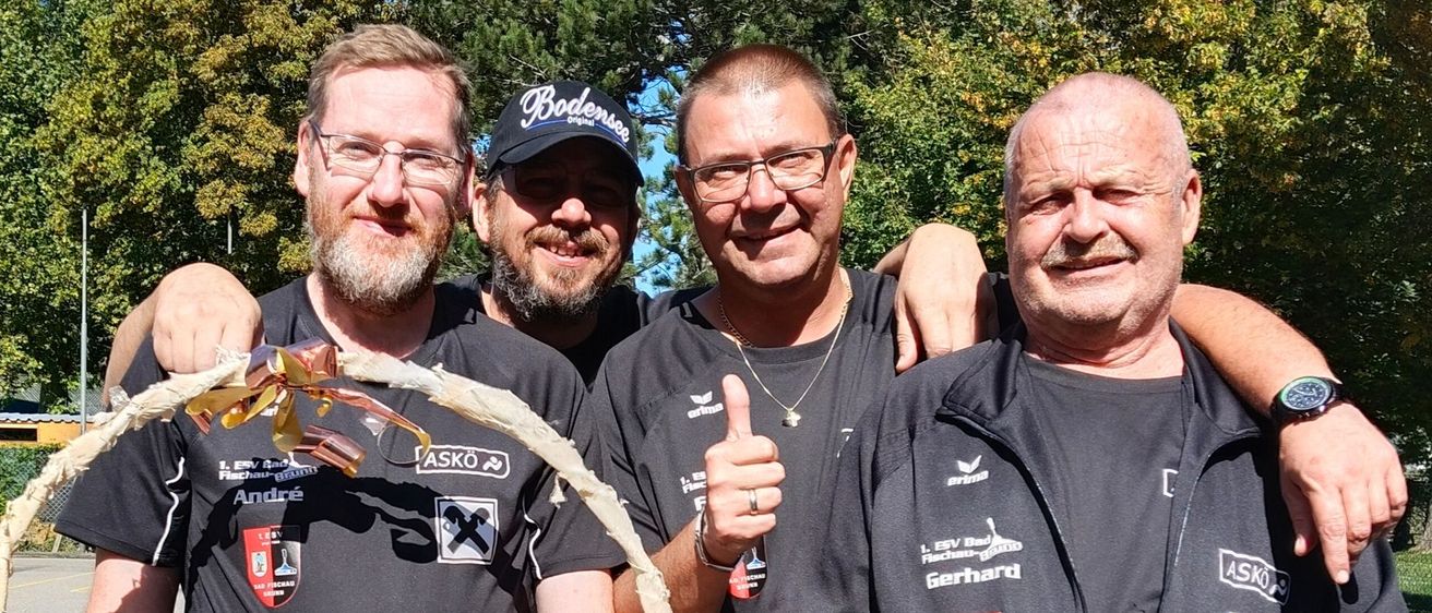 Vier Männer in schwarzer Sportbekleidung halten einen Korb voller Snacks. Ein Mann trägt eine Kappe und eine Brille, ein anderer zeigt den Daumen hoch. Sie stehen in einem Außenbereich mit Bäumen und einer Wasserrutsche im Hintergrund.