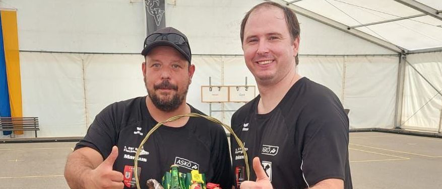 Zwei Männer in schwarzen Shirts und Shorts posieren mit einem Korb voller Süßigkeiten, lächeln und geben Daumen hoch in einem überdachten Bereich mit einem weißen Zelt.