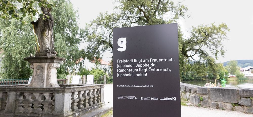 Schwarzer Informationsschild über Freistadt am Frauenteich, umgeben von Bäumen, mit einer Statue und einem Steinzaun im Hintergrund.