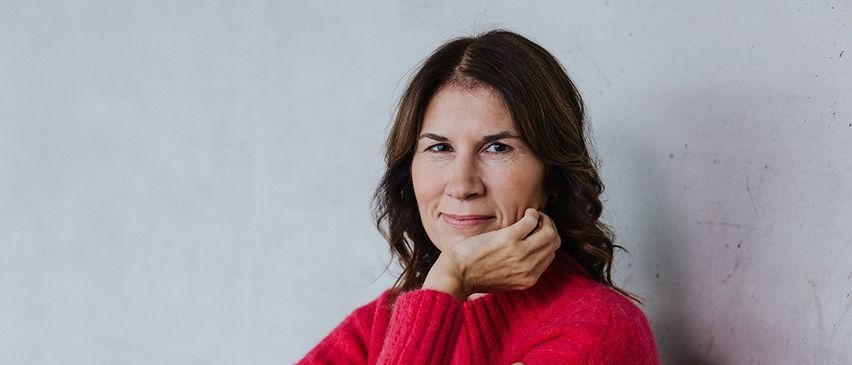 Eine Frau in einem roten Pullover und blauen Jeans sitzt mit verschränkten Armen an einer weißen Wand und blickt mit einem Lächeln zur Kamera.