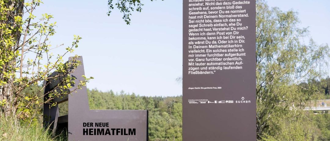 Ein Schild mit der Aufschrift "Der neue Heimatfilm" steht auf einem Grasweg. Dahinter befindet sich ein Baum mit der Nummer 9. Das Schild ist dunkel lila mit weißen Text. Der Hintergrund zeigt einen Grasbereich mit Bäumen.
