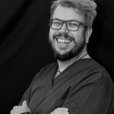 Ein schwarz-weißes Foto eines lächelnden Mannes mit Brille und Bart. Er trägt ein schwarzes Hemd und hat die Arme verschränkt.