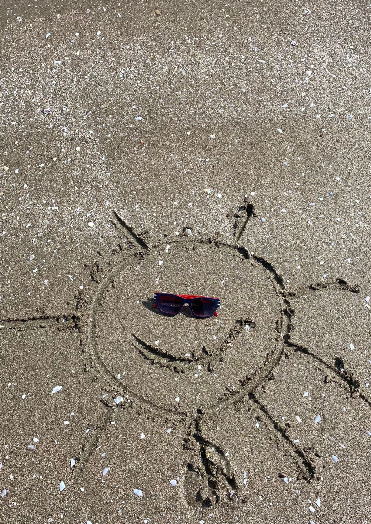 Ein in Sand gezeichneter Sonnenform mit einer Sonnenbrille darin, die einem lächelnden Gesicht auf einem Strand ähnelt.