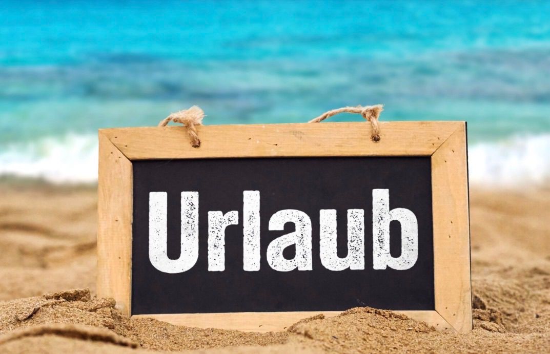 A blackboard with the word Urlaub written on it sits in the sand on a beach. The sea is calm and blue in the background.