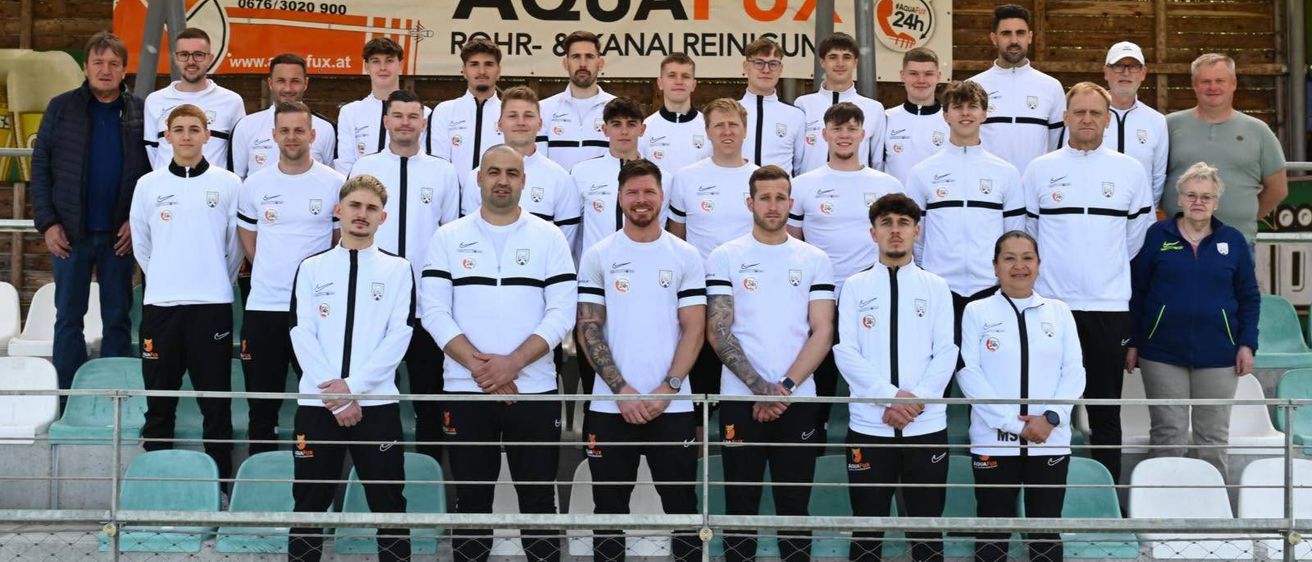 Eine Fußballmannschaft in weißen Trikots mit schwarzen Streifen steht vor einem Banner mit dem Text Aqua Fux. Die Spieler posieren für ein Gruppenfoto.