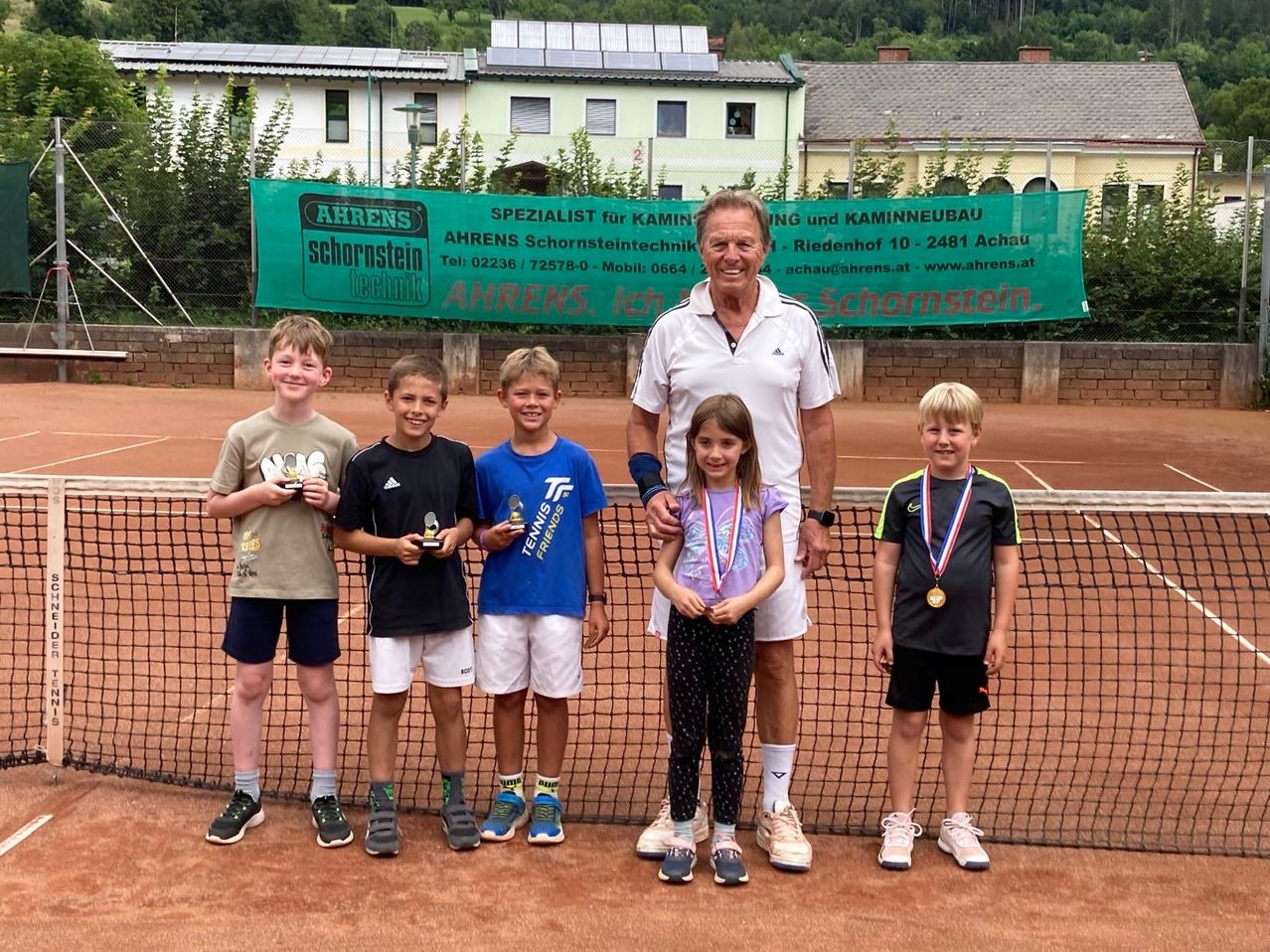 Eine Gruppe von Kindern und einem Erwachsenen stehen auf einem Tennisplatz mit einem Banner, das Ahrens Schornstein im Hintergrund liest. Die Kinder halten Medaillen.