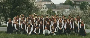 Ein Vintage-Foto einer Blaskapelle, beschriftet mit 'Zur Erinnerung', steht vor einer Kirche mit Turmspitze und fängt ein festliches Treffen im Jahr 1989 ein.