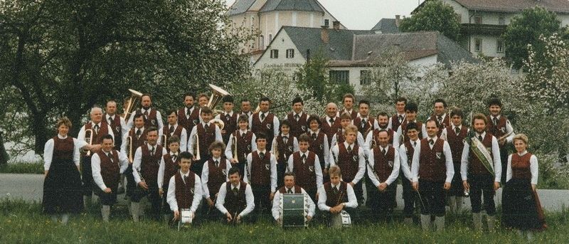 Ein Vintage-Foto einer Blaskapelle, beschriftet mit 'Zur Erinnerung', steht vor einer Kirche mit Turmspitze und fängt ein festliches Treffen im Jahr 1989 ein.