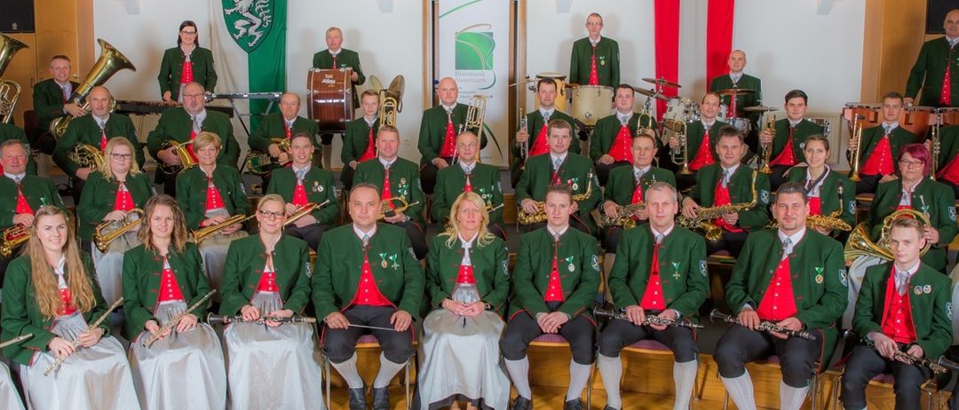 Eine Gruppe von Musikern, die grüne und rote Kleidung tragen, posiert für ein Foto. Sie halten verschiedene Musikinstrumente wie Trompeten, Posaunen, Trommeln und Flöten. Einige stehen, während andere auf Stühlen sitzen. Im Hintergrund ist ein grünes und weißes Schild zu sehen.