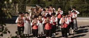 Eine Band spielt Instrumente auf einer Plattform über einer ländlichen Landschaft. Die Musiker tragen traditionelle Kleidung. Dahinter sind Bäume und eine entfernte Stadt sichtbar.