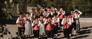 A band plays instruments on a platform above a rural landscape. The musicians wear traditional attire. Behind them, trees and a distant town are visible.