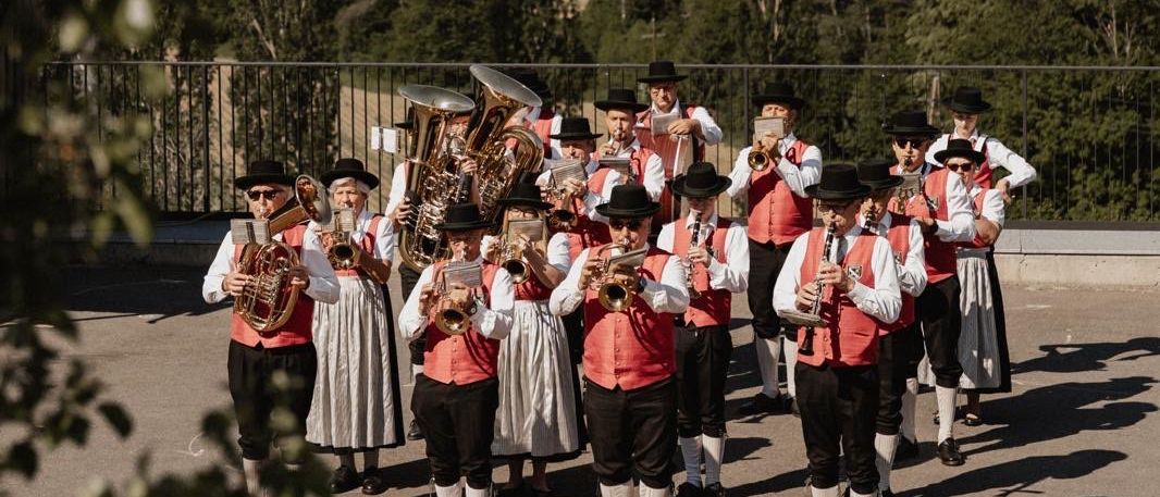 Eine Band spielt Instrumente auf einer Plattform über einer ländlichen Landschaft. Die Musiker tragen traditionelle Kleidung. Dahinter sind Bäume und eine entfernte Stadt sichtbar.