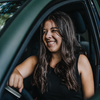 A woman with long brown hair is smiling while driving a car. She is wearing a black sleeveless shirt and gray pants.