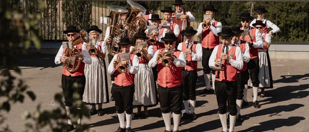 Eine Musikgruppe steht auf einer Terrasse und spielt verschiedene Instrumente unter einem klaren Himmel, mit einer ländlichen Landschaft im Hintergrund.