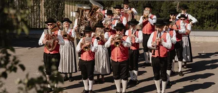 Eine Musikgruppe steht auf einer Terrasse und spielt verschiedene Instrumente unter einem klaren Himmel, mit einer ländlichen Landschaft im Hintergrund.