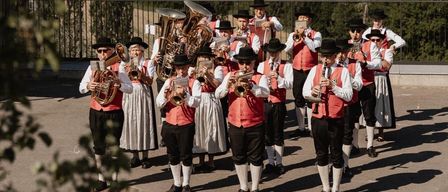 Eine Musikgruppe steht auf einer Terrasse und spielt verschiedene Instrumente unter einem klaren Himmel, mit einer ländlichen Landschaft im Hintergrund.