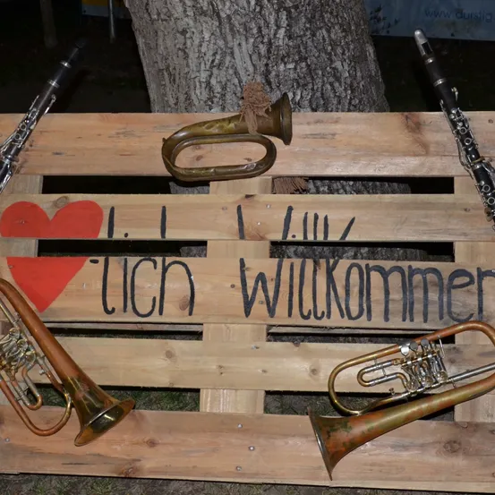 Eine Holzkiste zeigt Musikinstrumente wie Trompeten, ein Tenorhorn und eine Klarinette. Ein rotes Herz und deutsche Worte 'Ich willkomner' sind auf der Kiste.