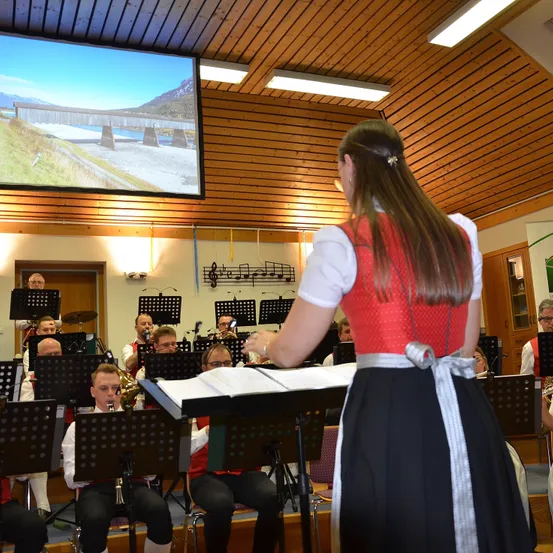 Eine Frau in traditioneller Kleidung steht vor einem Notenständer und dirigiert ein Blasorchester in einem Raum mit Holzwänden und einer Projektionsleinwand.
