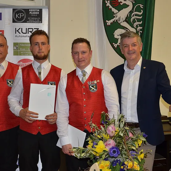 Vier Männer stehen zusammen, zwei tragen rote Westen mit Medaillen, einer hält ein Papier, und ein Mann im Anzug mit einem Blumenstrauß. Ein Banner mit einem Logo und Text ist hinter ihnen.