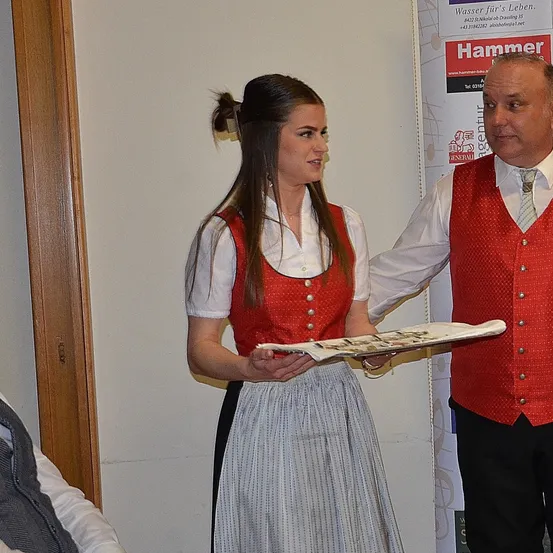 Eine Frau und ein Mann in traditioneller Kleidung stehen nebeneinander vor einer weißen Wand. Die Frau hält einen Tablett und der Mann trägt einen roten Westen mit Knöpfen.
