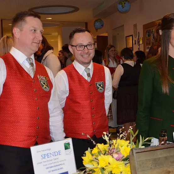 Zwei Männer in roten Westen stehen mit einer Frau in Grün in einem Flur. Sie haben Blumen und ein Schild, das Spende sagt. Andere Menschen sind im Hintergrund.