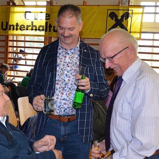 Zwei Männer stehen und lächeln mit Getränken in einem Raum mit einem Banner, das 'Cafe Unterhaltung' steht. Ein Mann hält eine grüne Flasche und der andere hat ein Glas. Andere Leute sitzen auf Stühlen hinter ihnen.