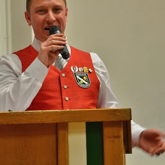 Ein Mann steht hinter einem Podium, hält ein Mikrofon in der Hand und trägt eine rote Weste mit weißen Punkten und einem Logo. Er scheint eine Rede zu halten.