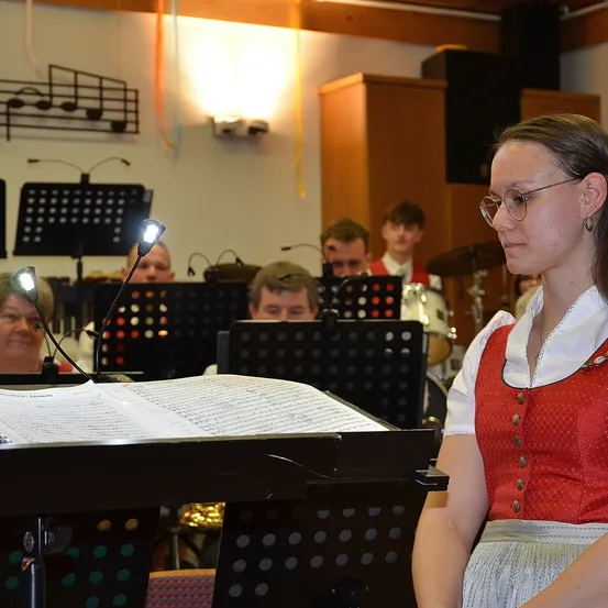 Eine Frau in traditioneller bayerischer Kleidung steht bei einem Notenständer mit einem Buch mit Noten. Dahinter spielt eine Band mit Trommeln und Blechblasinstrumenten.