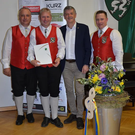 Vier Männer stehen zusammen in einem Raum. Zwei von ihnen tragen rote Westen und weiße Langarmhemden, während die anderen Anzüge tragen. Sie halten alle eine Urkunde. Vor ihnen steht eine Blumenvase.