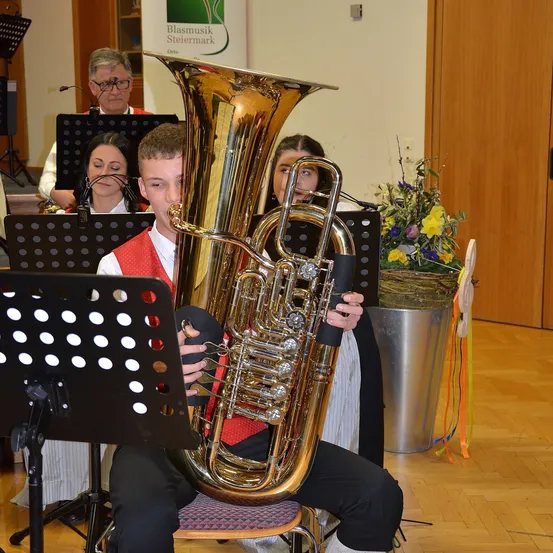 Ein junger Mann spielt ein blasinstrument, während er auf einem Stuhl in einem Raum mit musikalischen Instrumenten sitzt. Hinter ihm spielen zwei Frauen und ein Mann ebenfalls Instrumente. Rechts steht ein Blumenarrangement.