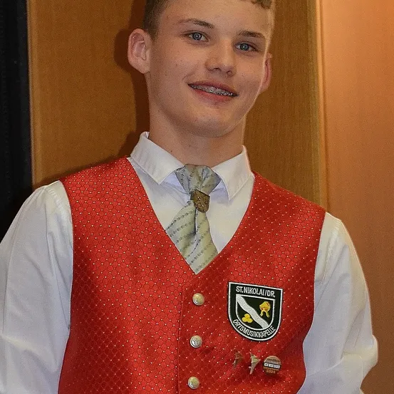 Ein junger Mann mit Zahnspange steht drinnen und trägt ein rotes Weste über einem weißen Hemd und Krawatte. Die Weste hat ein Logo und Abzeichen. Er lächelt.
